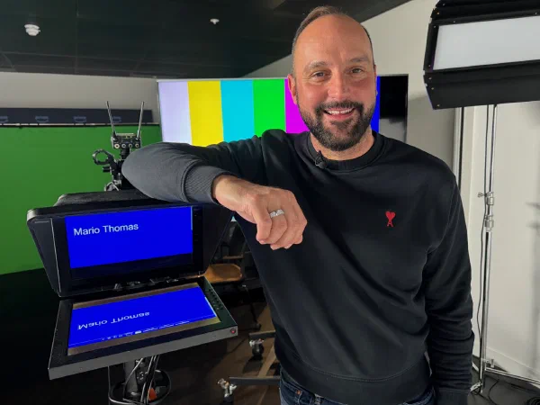 Mario Thomas in a broadcast studio in London beside a teleprompter showing his name