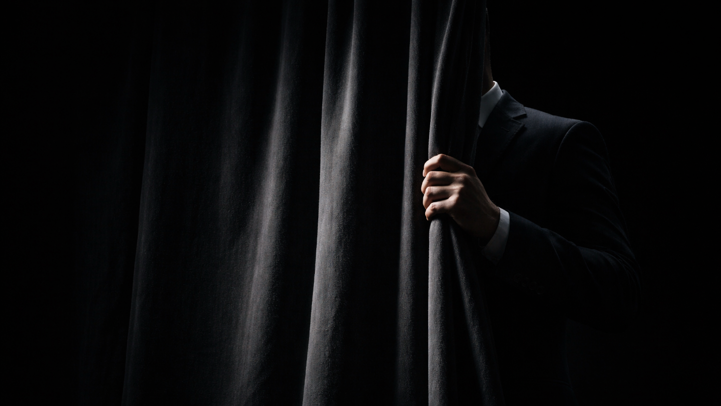 A figure in a dark suit, partially concealed behind a heavy charcoal velvet curtain, one hand gripping the curtain edge in sharp directional light against a black background — a visual metaphor for the unseen operator whose workings a director is expected to trust without seeing.