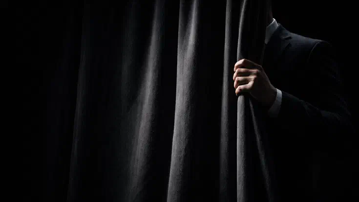 A figure in a dark suit, partially concealed behind a heavy charcoal velvet curtain, one hand gripping the curtain edge in sharp directional light against a black background — a visual metaphor for the unseen operator whose workings a director is expected to trust without seeing. (Image generated by ChatGPT 5.4)