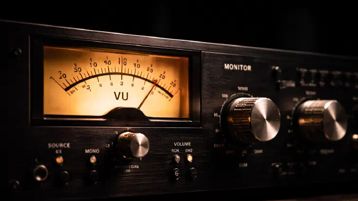 A close-up photograph of a professional audio mastering console, showing a warmly lit analogue VU meter on the left with its amber-glowing face, flanked by precision control knobs and monitoring switches on a dark panel. The shallow depth of field draws the eye to the meter itself, with the surrounding controls falling gently into shadow. An image representing the precision instruments used by audio engineers to measure fidelity, used here as a metaphor for the four indicator types that give Boards maximum fidelity on the decisions in front of them (Image generated by ChatGPT 5.4)