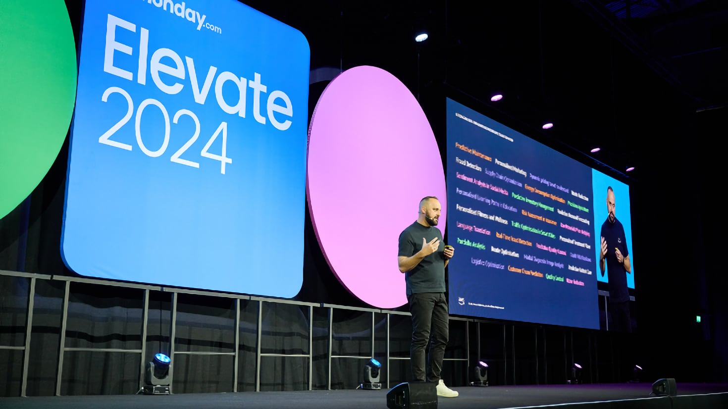Mario Thomas on stage at Monday.com Elevate at ExCel London with a list of AI use cases on screen.