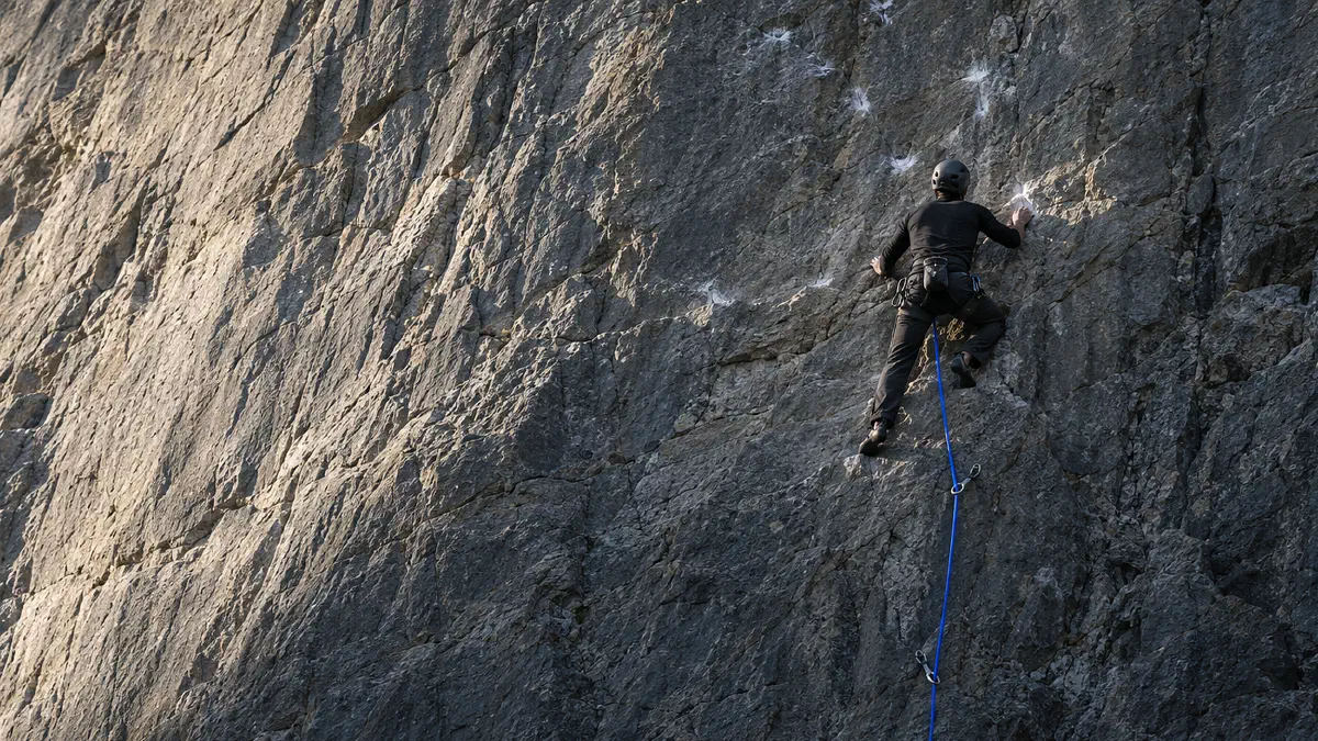 A climber seen from behind pauses high on a grey rock face, reading the rock above, an electric-blue rope tracing the route already climbed below them: a strategy is the line up the whole face, not the next hold. (Image generated by ChatGPT 5)